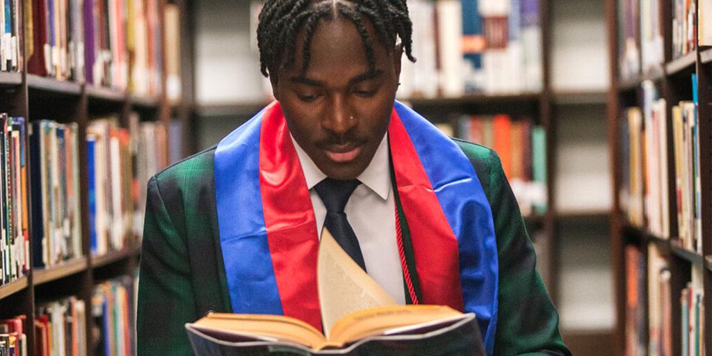 A person wearing a graduation stole reads a book in a library aisle.